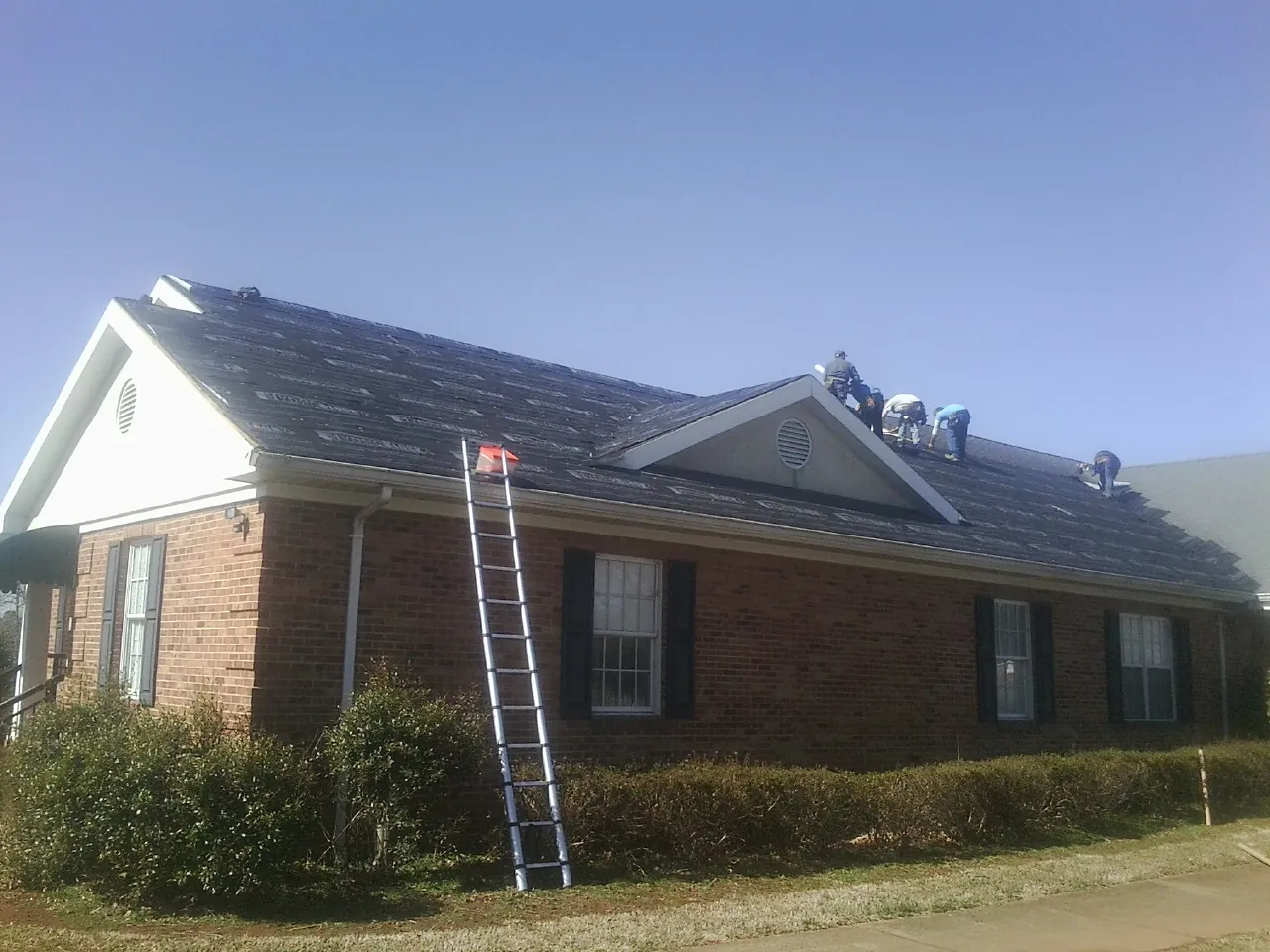 Crew tearing shingles off a brick ranch roof with a ladder set — Metal Roof Repair work by our crew in Woodman
