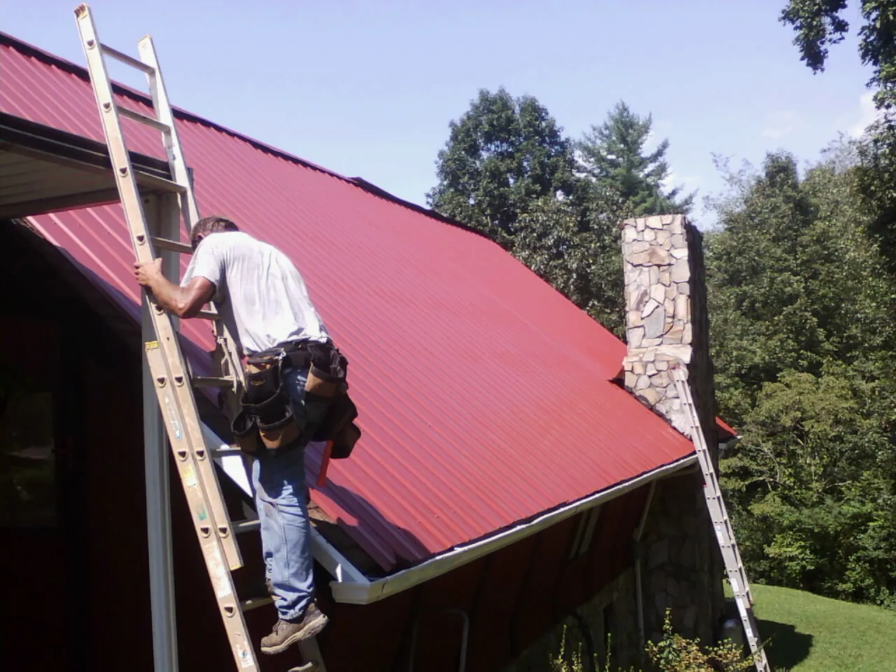 Roofer on a ladder beside a red metal roof and stone chimney — Soffit & Fascia Repair by Roofing Brothers in Woodman